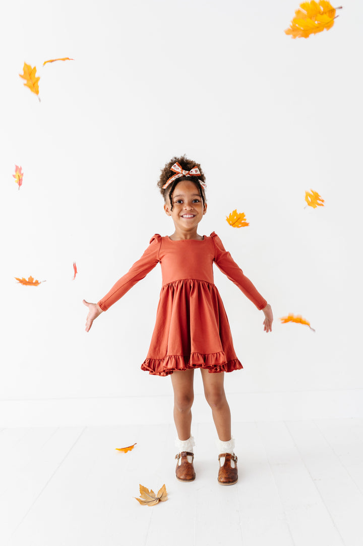 little girl throwing fall leaves in rust long sleeve twirl dress paired with ivory lace bobby socks and rust floral hair bow