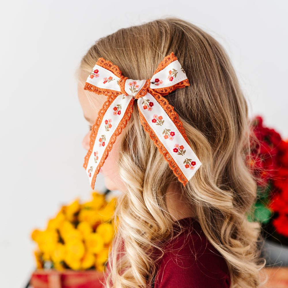 young girl by fall flowers in cream bow with rust floral pattern and rust lace trim paired with burgundy twirl dress