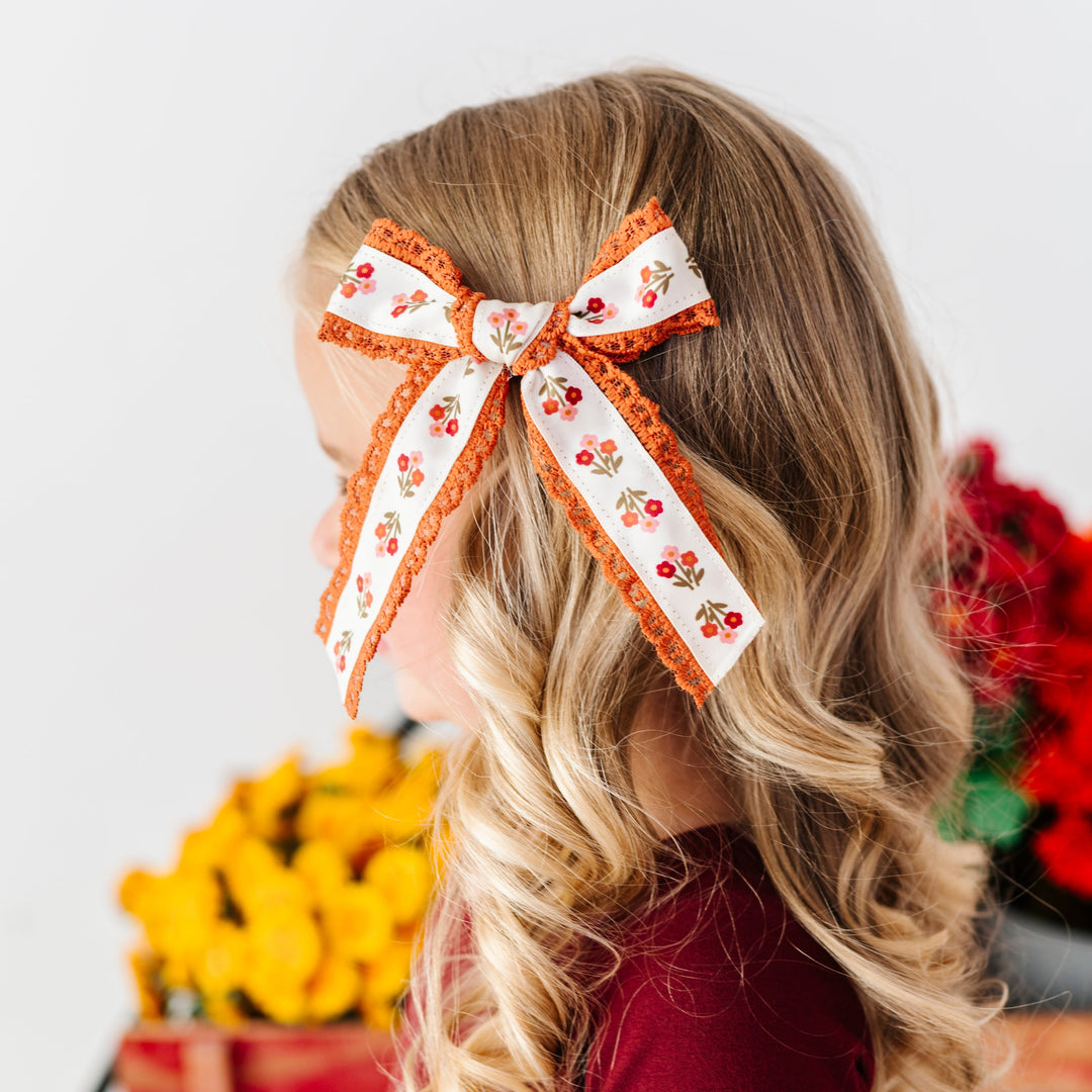 young girl by fall flowers in cream bow with rust floral pattern and rust lace trim paired with burgundy twirl dress