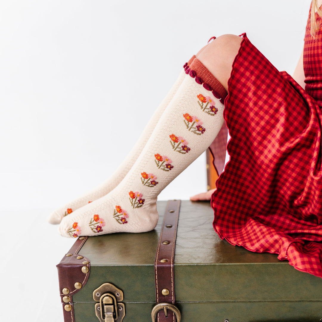 girl sitting on green vintage suitcase wearing cream knee socks with fall flower bouquet pattern and rust trim paired with rust gingham dress