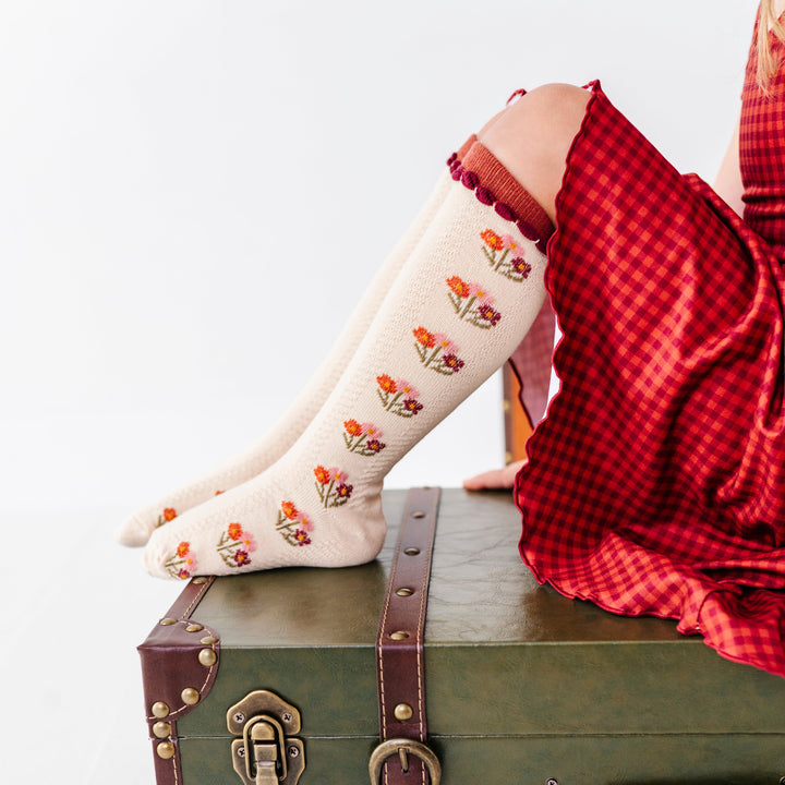 girl sitting on green vintage suitcase wearing cream knee socks with fall flower bouquet pattern and rust trim paired with rust gingham dress