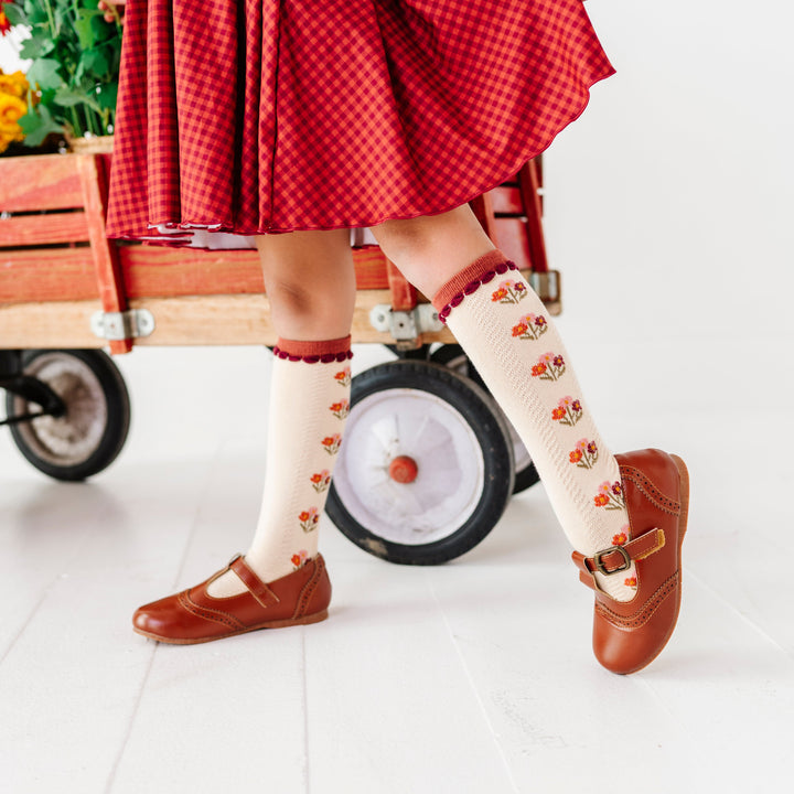 girl wearing brown mary janes and rust gingham fall dress paired with cream knee socks with fall floral design and rust trim