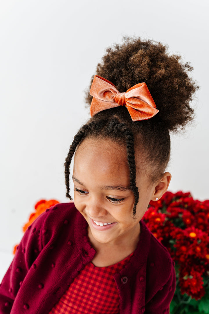 young girl with rust velvet hair bow clipped to ponytail paired with rust gingham dress and coordinating burgundy cardigan