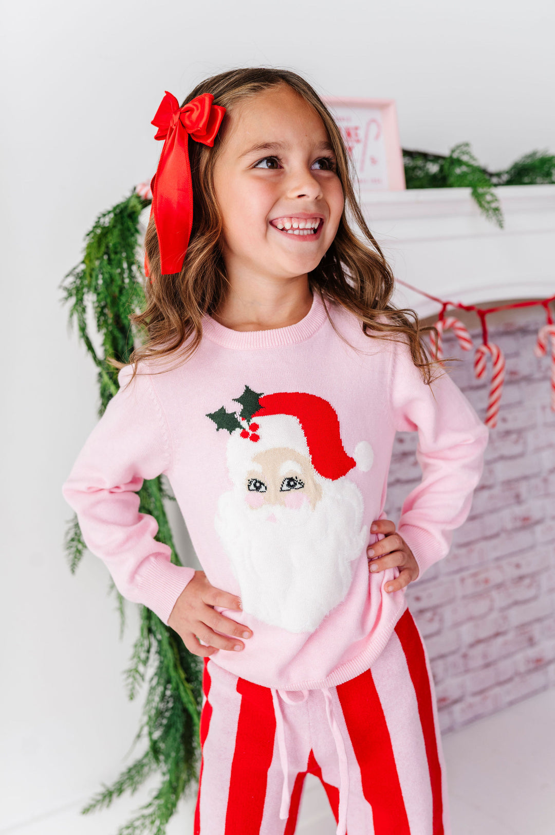 A young girl smiling and looking to the side, wearing a light pink long-sleeve pajama top with a large Santa Claus face graphic on the front, paired with red and white striped pajama pants. She has a large red bow in her hair, standing in front of a decorated holiday fireplace background.