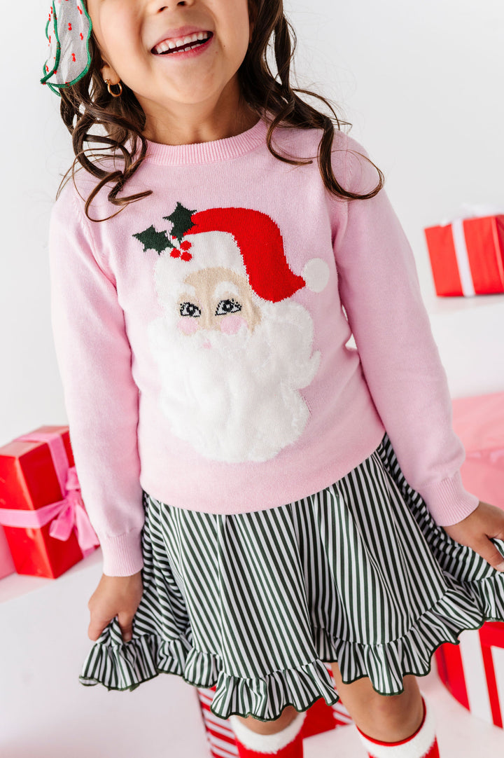 A young girl wearing a pink long-sleeve sweater with a large Santa Claus face and hat graphic on the front, paired with a black and white striped ruffled skirt. She is standing in a festive setting with red wrapped gift boxes in the background.