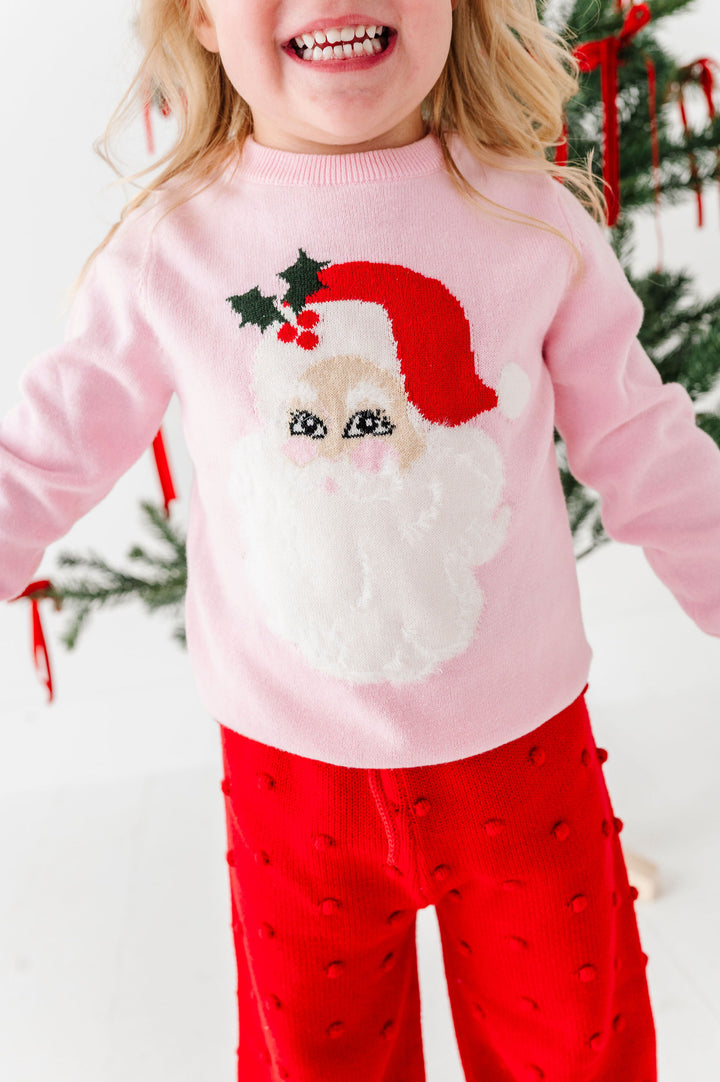 A young child wearing a light pink knit sweater with a large knitted Santa face and red hat, paired with bright red pants, standing in front of a decorated Christmas tree.