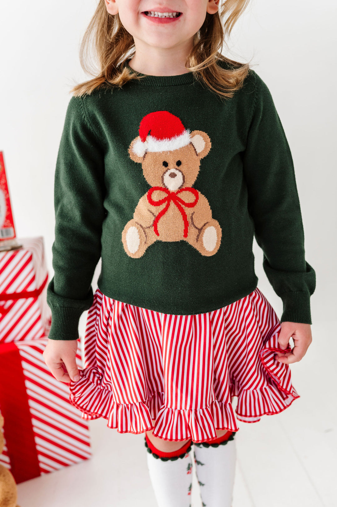 A young child wearing a dark green knit sweater with a teddy bear graphic in a Santa hat and red bow, paired with a red and white striped ruffled skirt, standing in a holiday-themed setting.