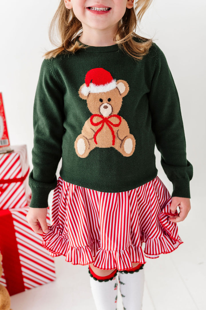 A young child wearing a dark green knit sweater with a teddy bear graphic in a Santa hat and red bow, paired with a red and white striped ruffled skirt, standing in a holiday-themed setting.