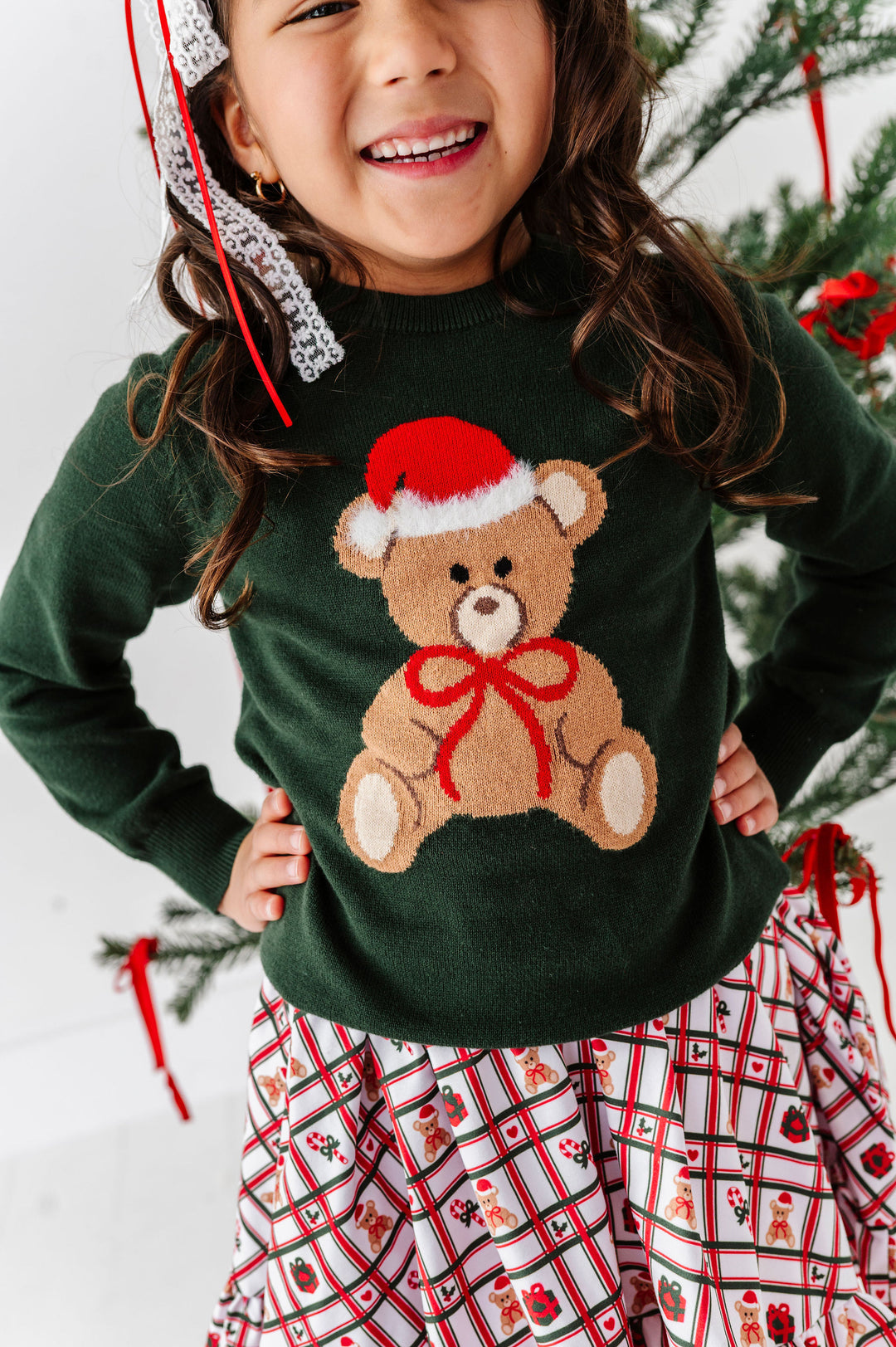 A smiling young girl wearing a dark green knit sweater with a large teddy bear graphic wearing a Santa hat and red bow, paired with a red, white, and black plaid skirt. She is standing in front of a decorated Christmas tree.