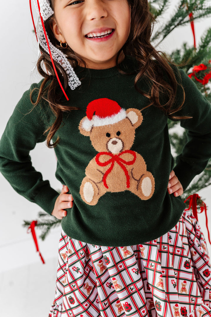 A smiling young girl wearing a dark green knit sweater with a large teddy bear graphic wearing a Santa hat and red bow, paired with a red, white, and black plaid skirt. She is standing in front of a decorated Christmas tree.
