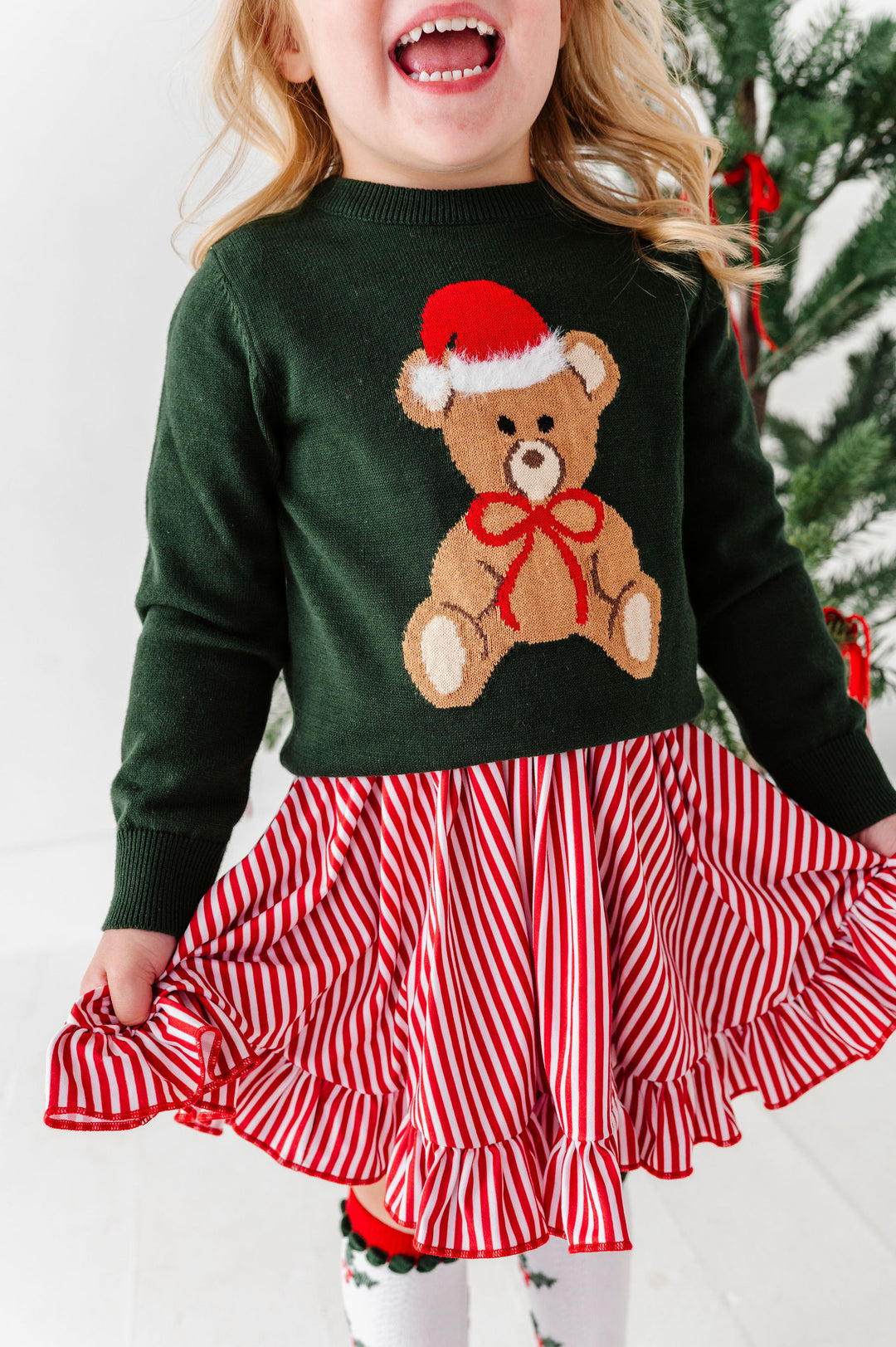 A young child wearing a festive Christmas sweater and skirt set. The dark green knit sweater features a large brown teddy bear graphic wearing a red Santa hat and red bow.