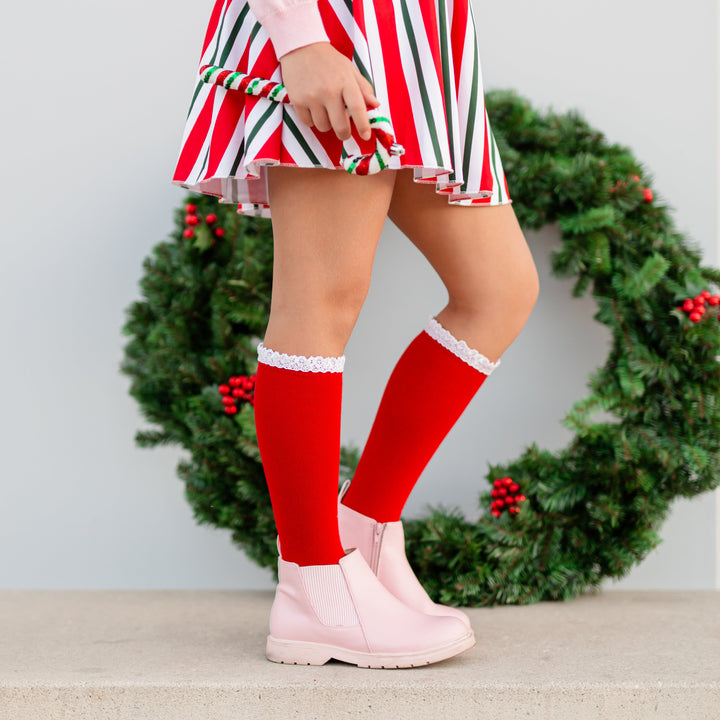 girl in front of Christmas wreath in red knee high socks with white lace trim paired with candy cane striped skirt