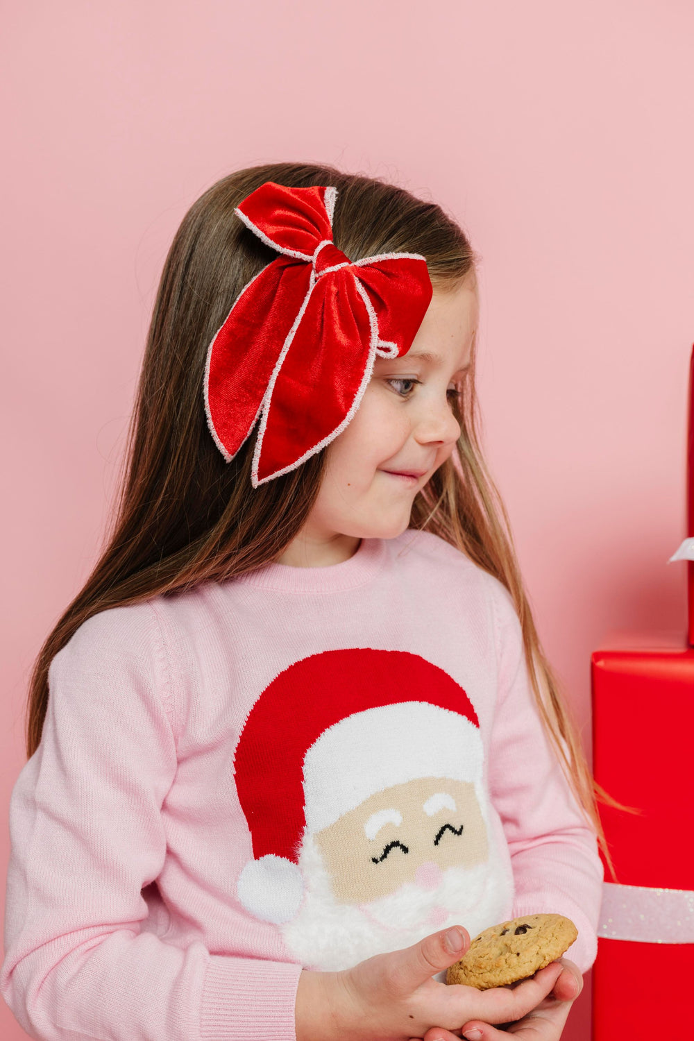 little girl holding cookie in pink Santa sweater wearing bright red velvet bow with white velvet trim