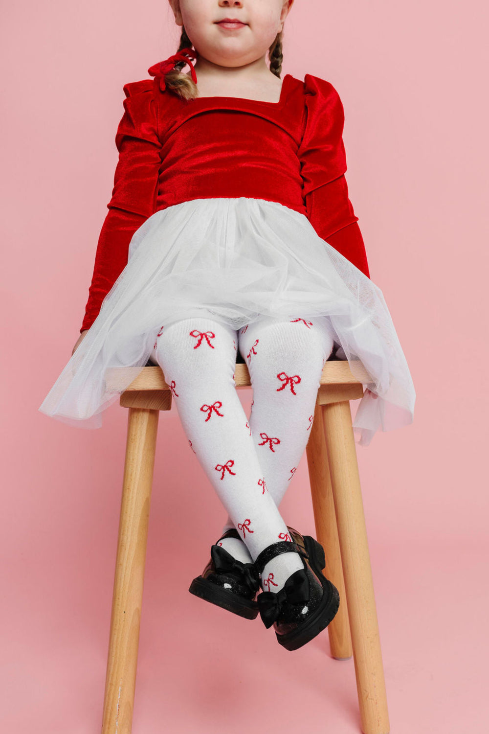 young girl sitting in red velvet, white tutu Christmas dress paired with white tights with red bow design and black mary jane shoes