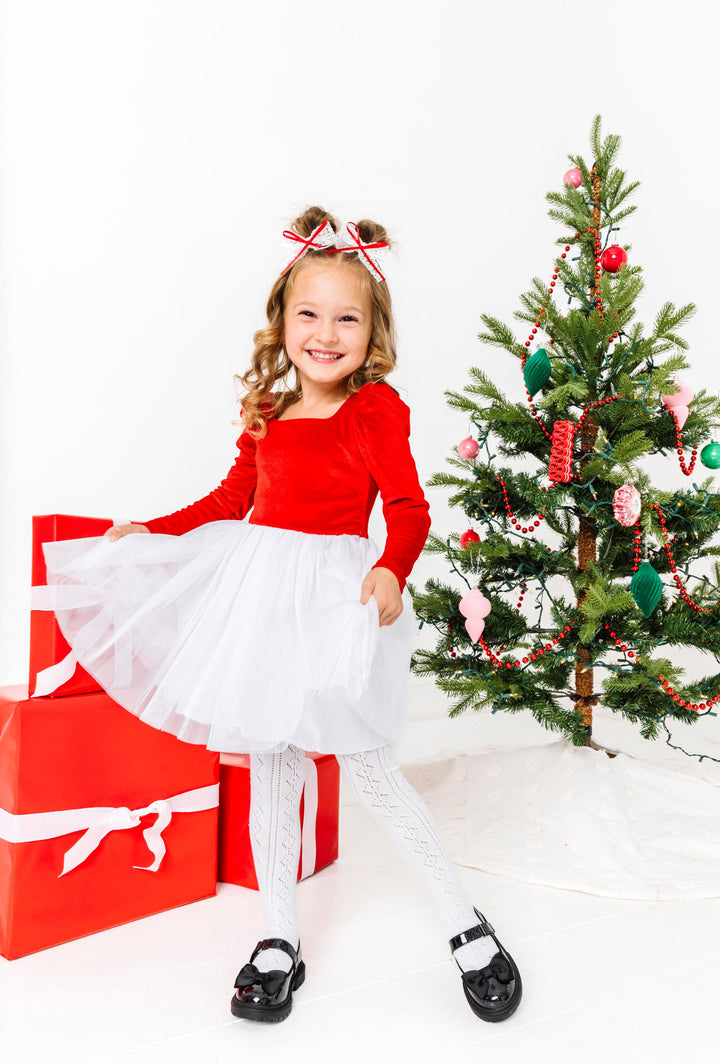little girl by Christmas tree wearing Santa inspired tutu dress with bright red long sleeve velvet bodice and white tulle skirt paired with matching lace pigtail bows and white open knit tights