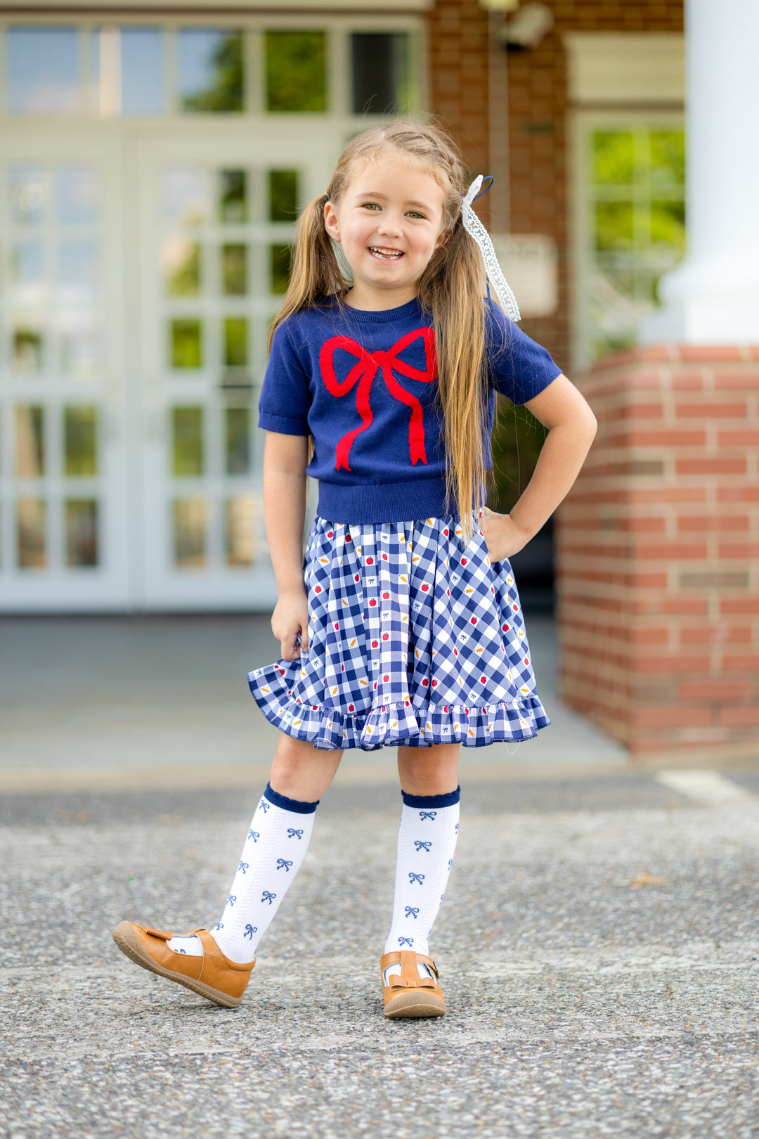 little girl at school with school themed navy gingham print twirl dress, short sleeved navy sweater with red bow design and blow pattern knee highs