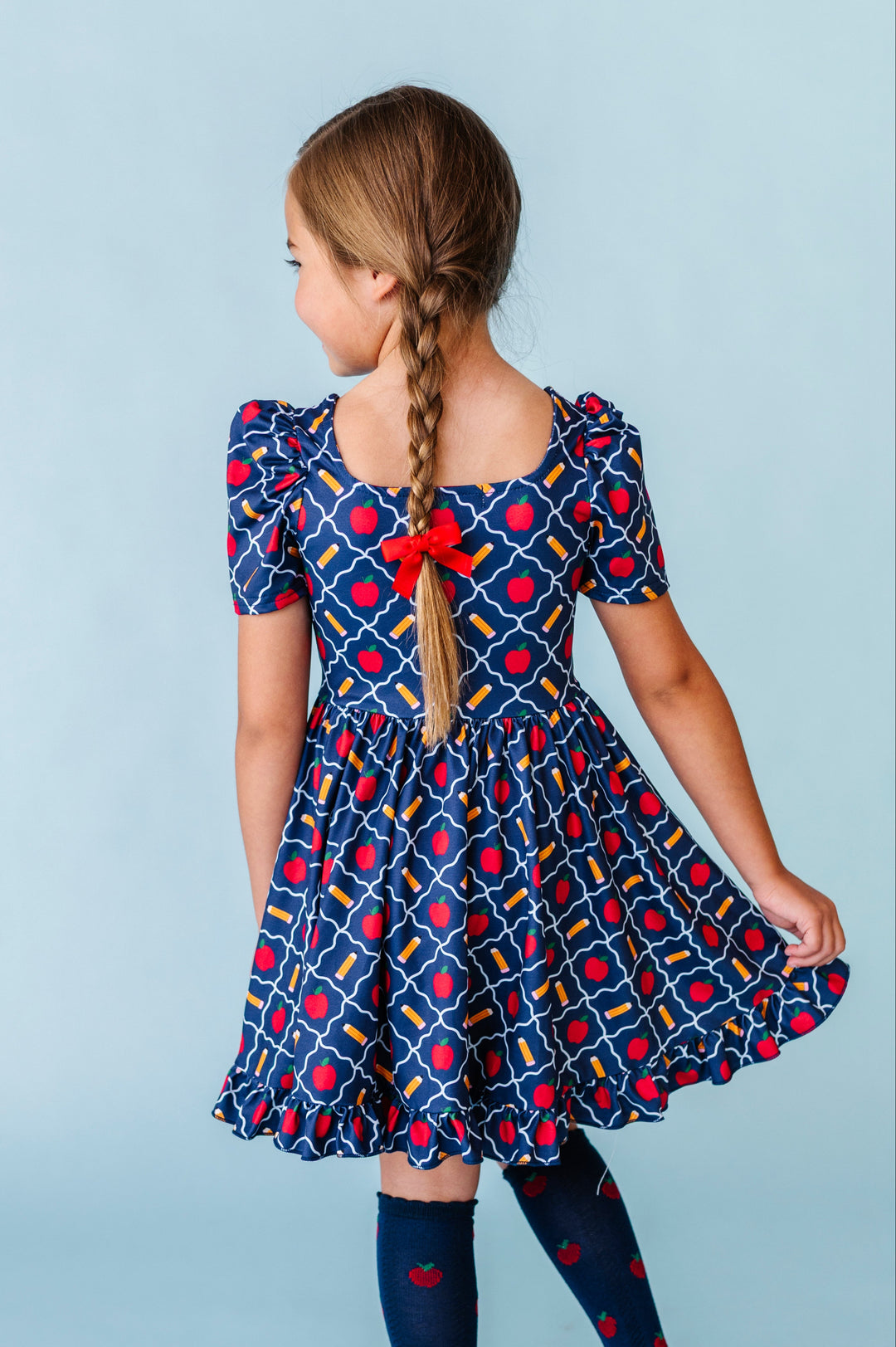 back view of little girl in braids wearing square neck navy school print twirl dress with matching red pigtail bows and apple knee socks