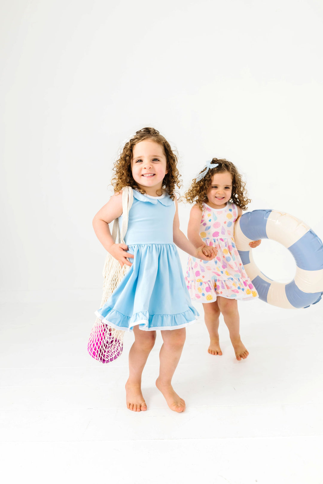 sisters holding beach toys in light blue and seashell print sleeveless twirl dresses for summer