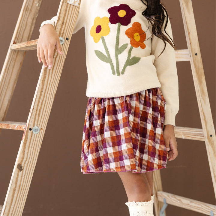 little girl standing by ladder in cream knit sweater with fall flower design and matching burgundy and pumpkin velvet plaid skort
