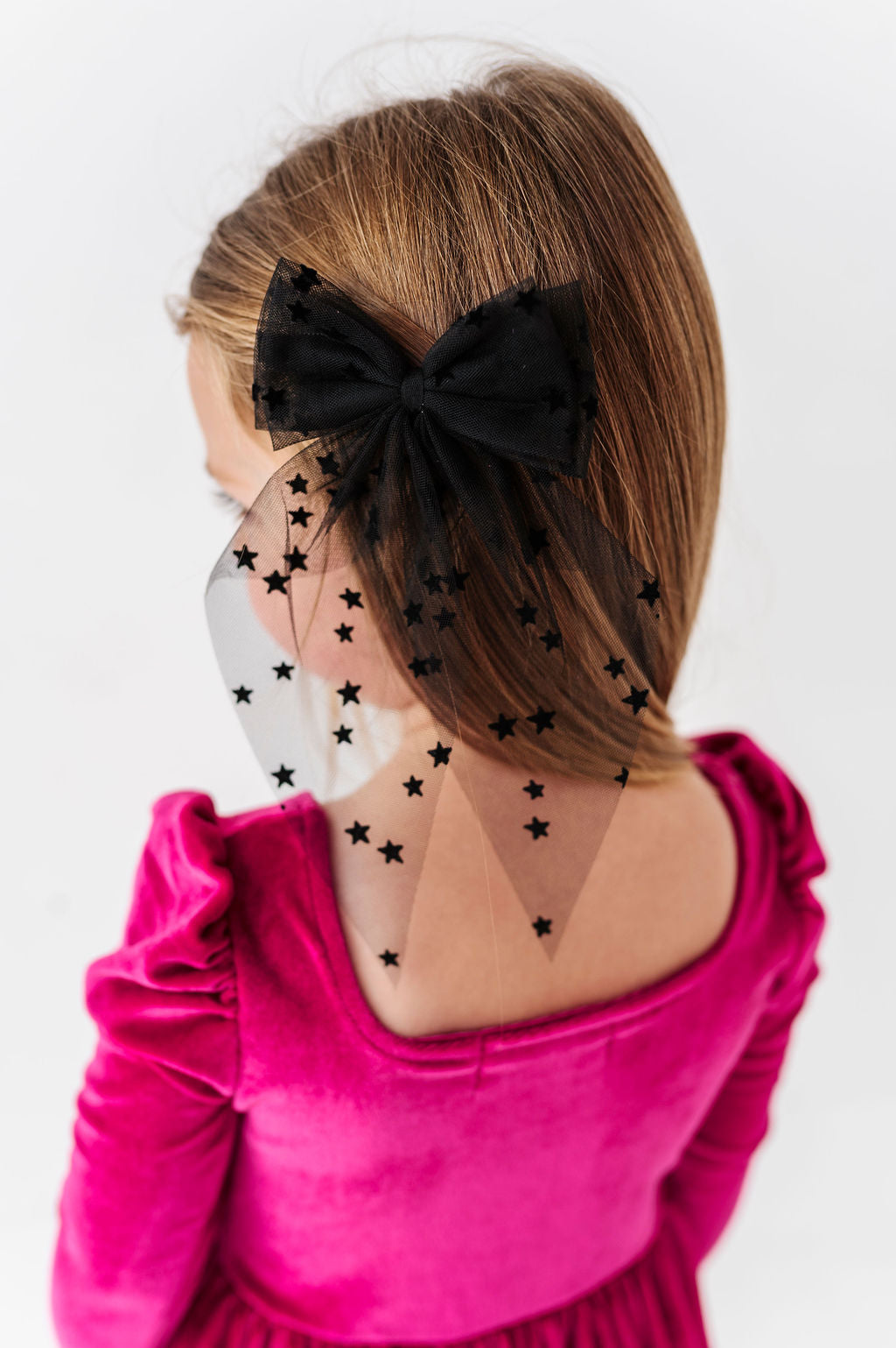 back view of little girl in square neck, long sleeve, hot pink velvet dress paired with sheer oversized black tulle hair bow with tiny velvet stars for Halloween