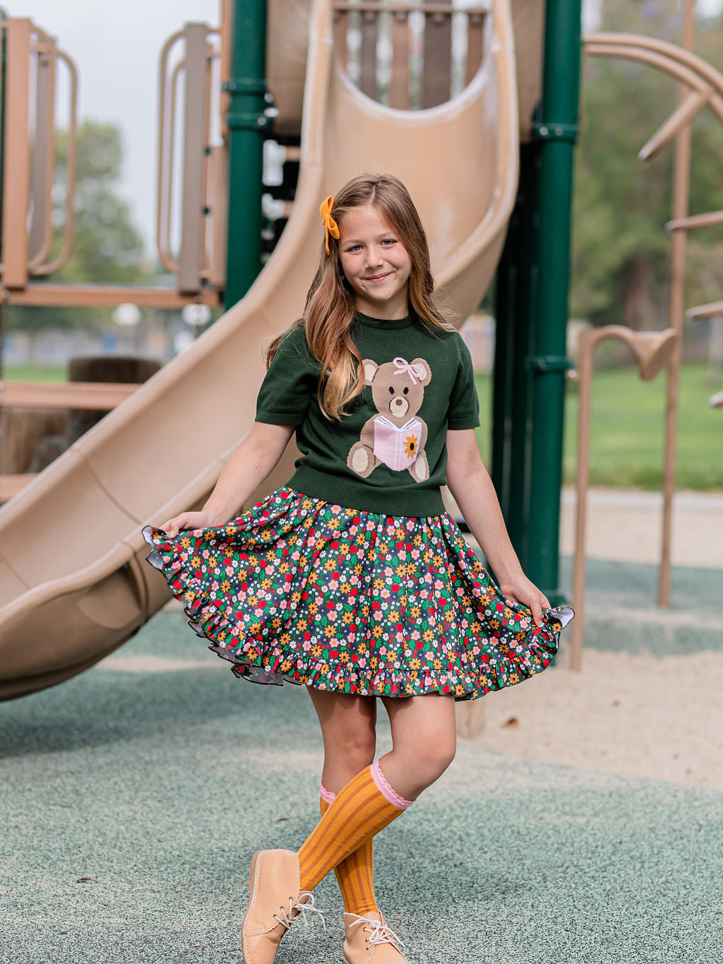 girl in front of playground slide in green bear sweater paired with green apple and flower print twirl and pencil themed knee socks