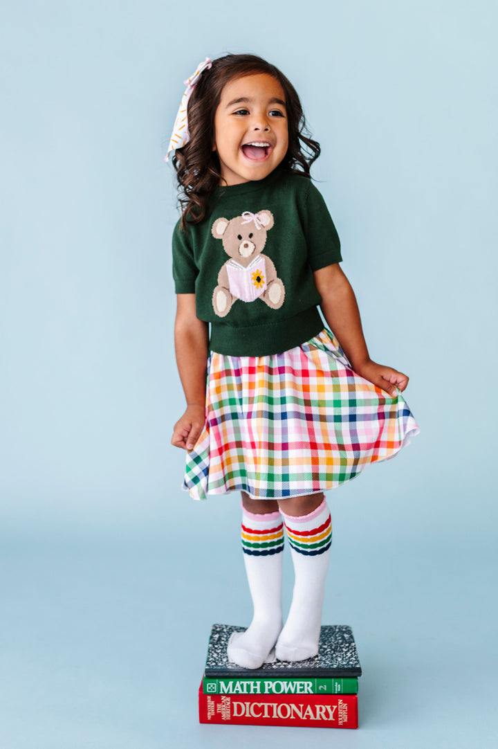 little girl standing on books in green short sleeve knit sweater with bear reading book design paired with primary gingham skort and matching striped knee socks