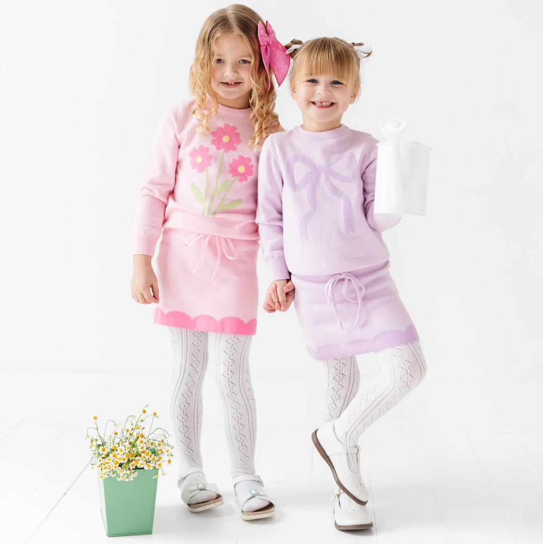 two girls holding hands wearing coordinating outfits- one is a pink cotton knit skirt and pink top with flowers and the other is a purple skirt with matching sweater on top with bow