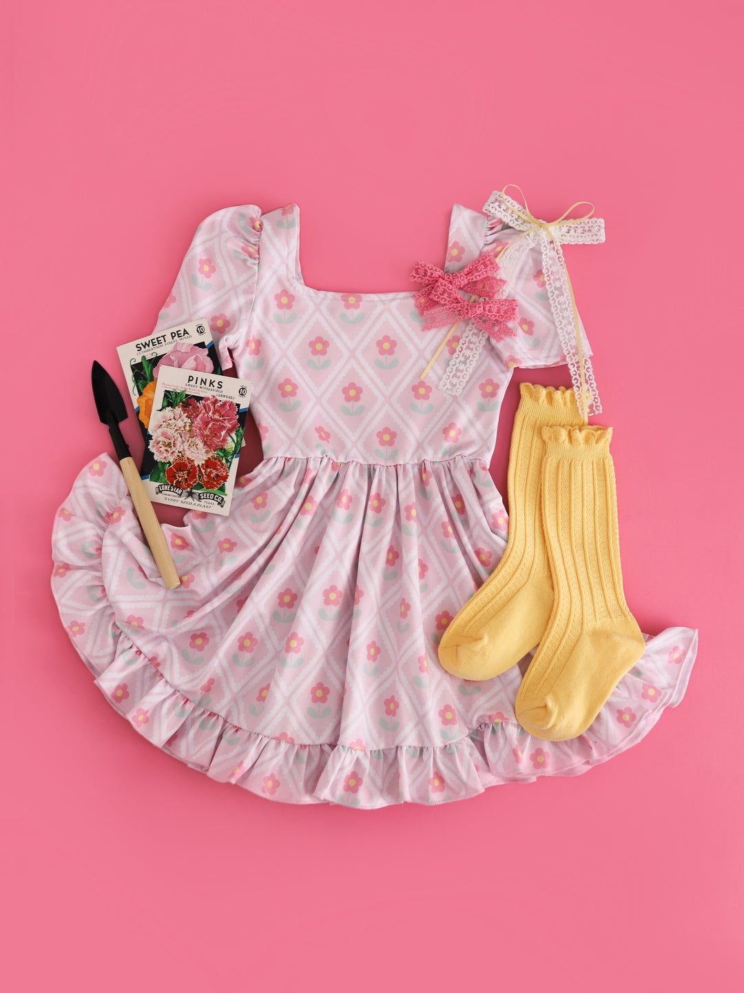 flat lay of girls' pastel pink square neck twirl dress with lattice-like scalloped design surrounding cute pink daisies styled with pastel yellow accessories 