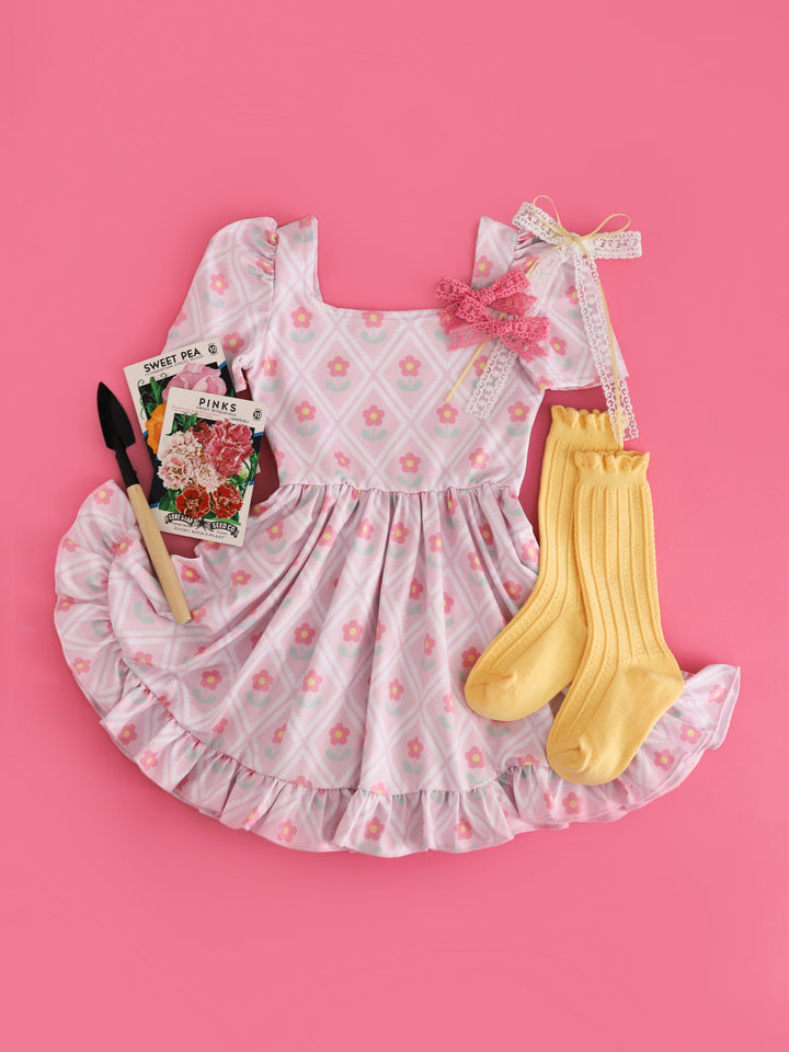 flat lay of girls' pastel pink square neck twirl dress with lattice-like scalloped design surrounding cute pink daisies styled with pastel yellow accessories 