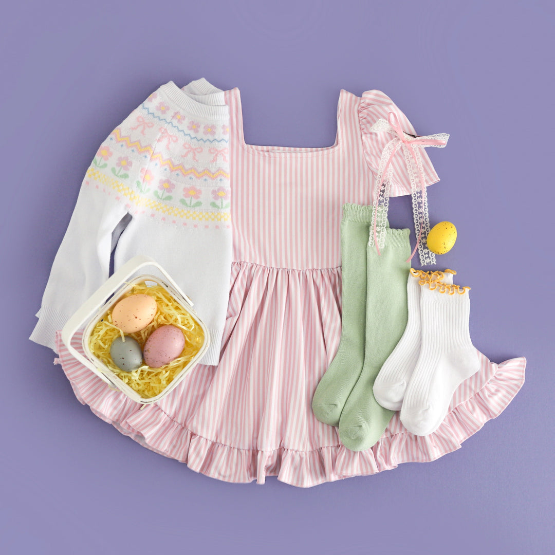 flat lay styled photo with easter egg basket featuring girls' pastel pink and white pinstripe twirl dress and white spring print fair isle sweater with accessories to match