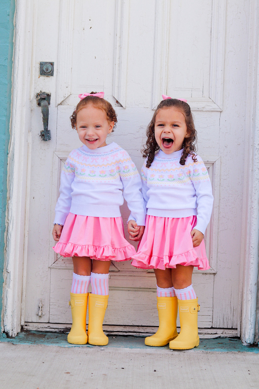 little girls in matching white with pastel fair isles pattern sweaters, light pink twirl dresses, matching pink and white vertical striped knee highs and yellow rain boots