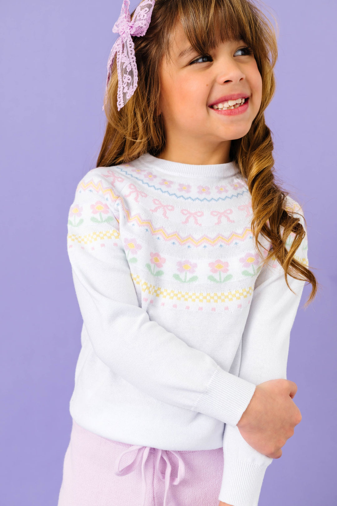 young girl in white cotton knit sweater with pastel spring fair isle print and matching lilac pants and lace bow