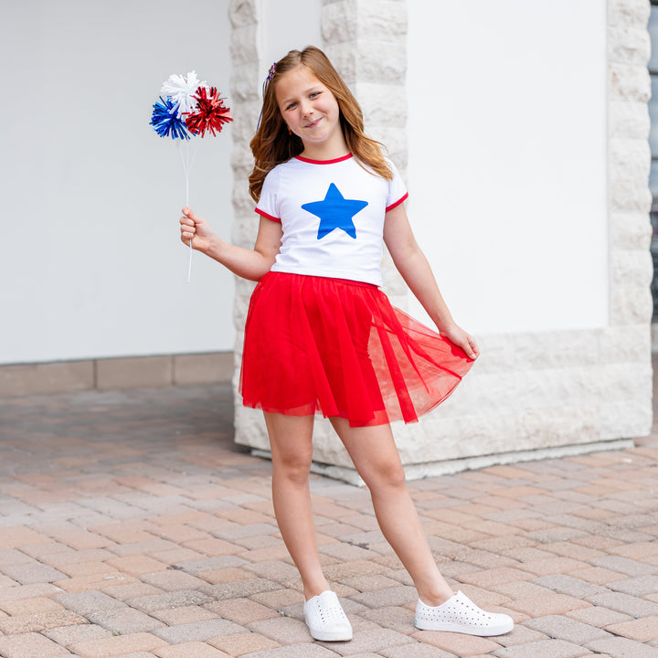 little girl holding red, white and blue pom poms in white fitted tee with blue star design and matching red tutu skirt with built in shorts for 4th of July parade