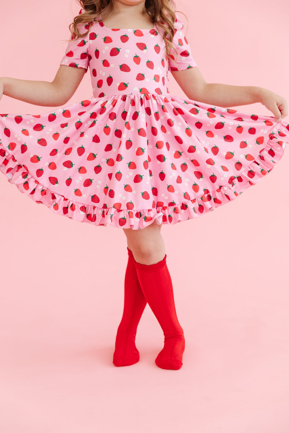girl wearing girls' pink short sleeve twirl dress with red strawberry print and dainty white flowers and matching red knee high socks
