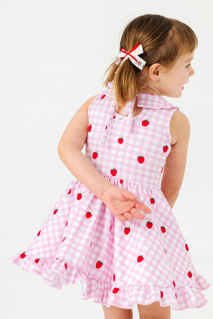 back view of young girl twirling in pink gingham and strawberry print summer twirl dress and matching white lace and red ribbon pigtail bows