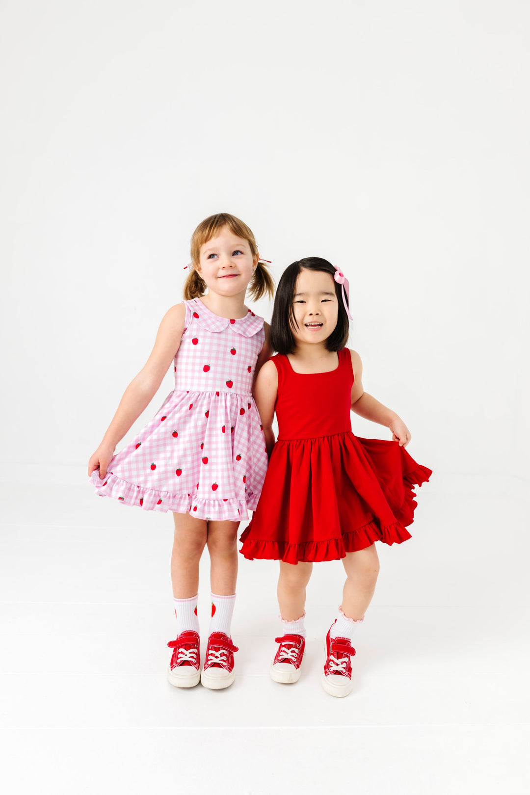 two girls in coordinating strawberry gingham and bright red sleeveless summer twirl dresses paired with matching midi socks and red sneakers