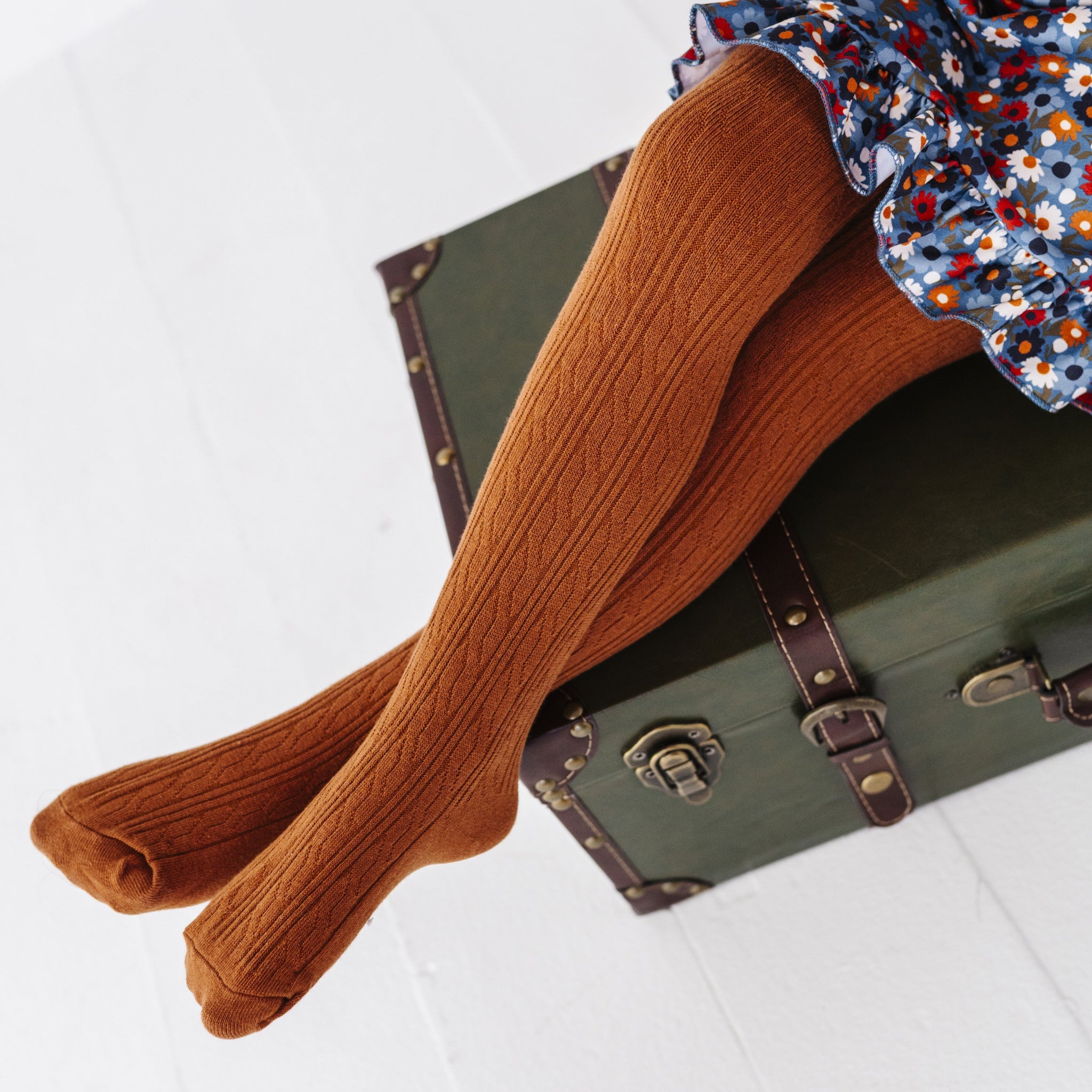 little girl sitting on vintage green suitcase wearing warm brown cable knit tights paired with coordinating blue floral twirl dress