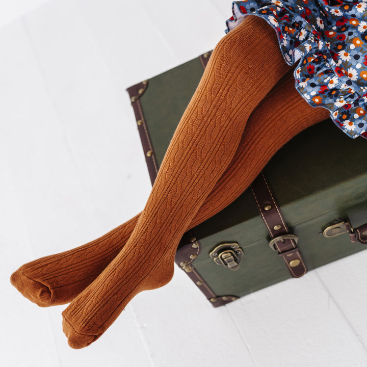 little girl sitting on vintage green suitcase wearing warm brown cable knit tights paired with coordinating blue floral twirl dress
