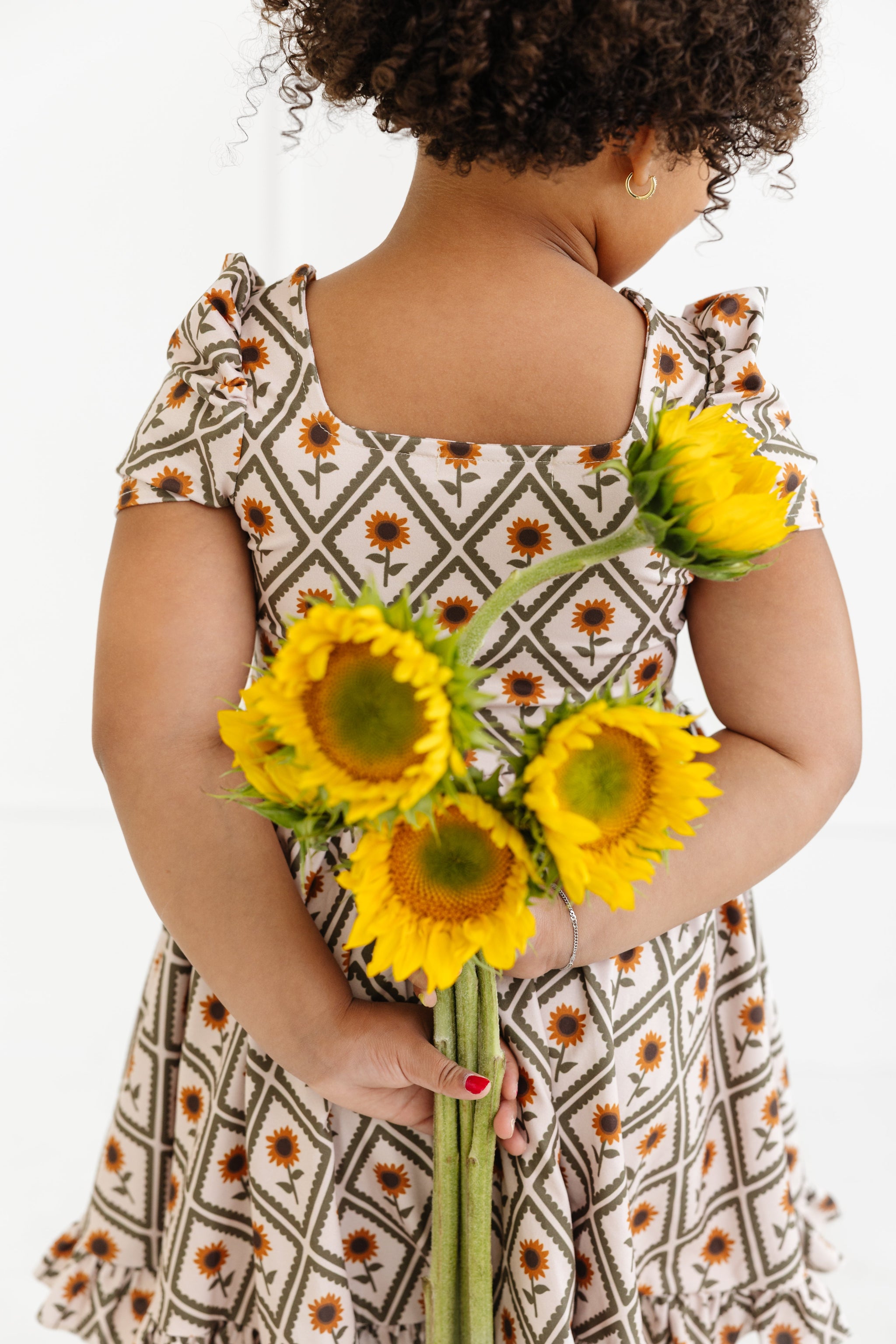 little girl holding sunflowers behind her back in cream square neck twirl dress with sunflowers in scalloped diamond pattern