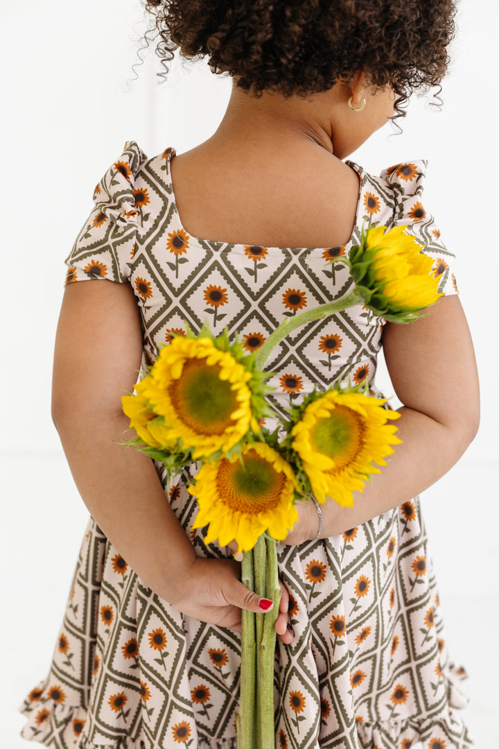 little girl holding sunflowers behind her back in cream square neck twirl dress with sunflowers in scalloped diamond pattern