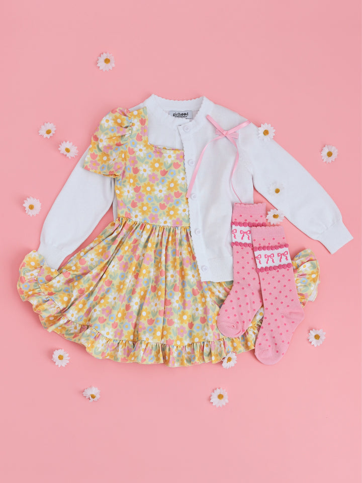 flatlay of little girls spring outfit with pastel yellow square neck twirl dress with white cardigan and pink knee high socks with bows and dots printed on them