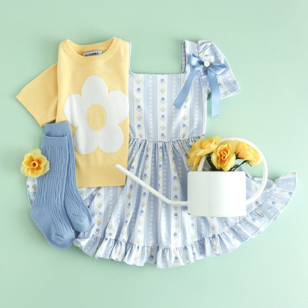 flat lay with watering can for girls' pastel blue spring wallpaper print twirl dress paired with light yellow short sleeve flower sweater and matching blue socks and bow