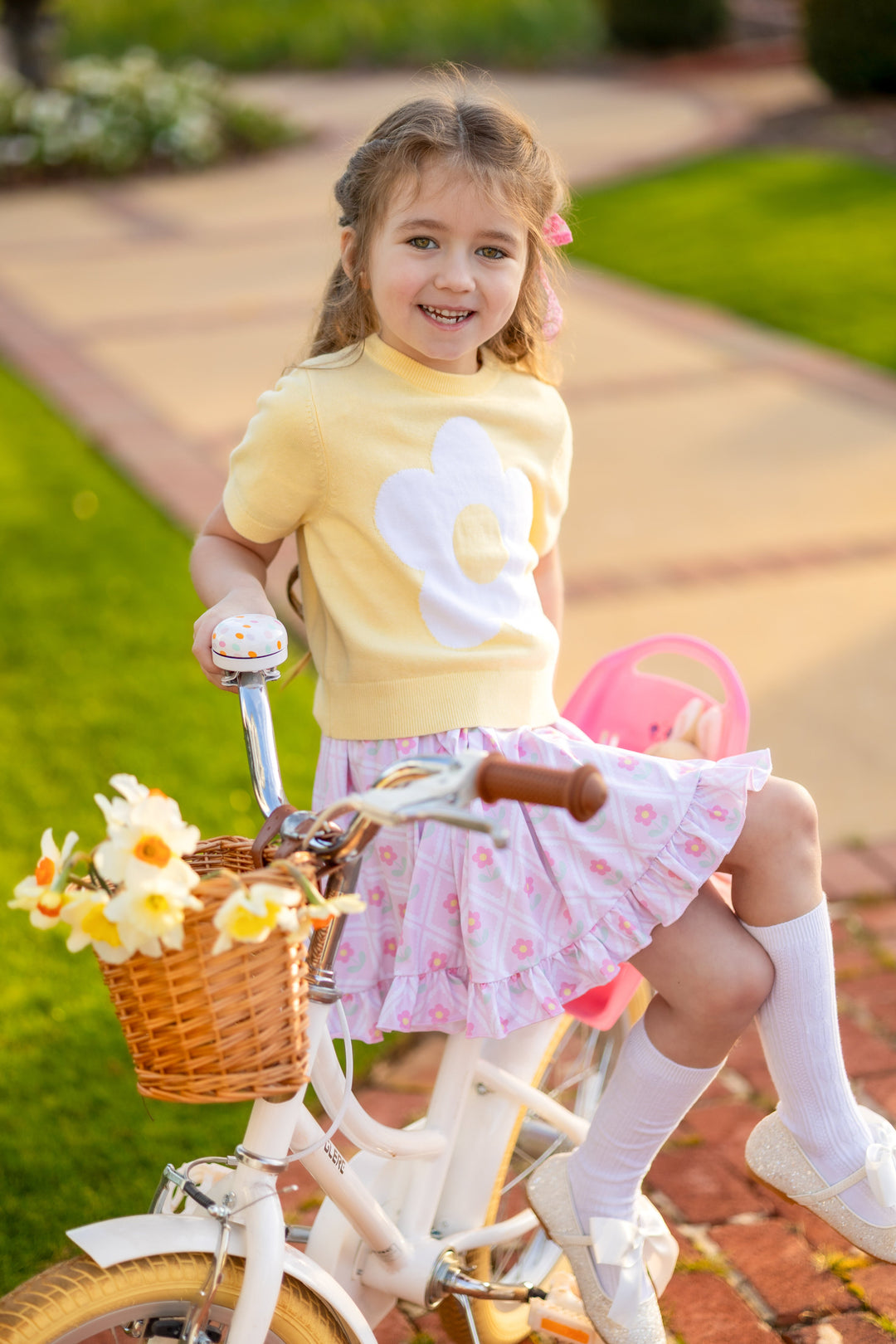 little girl sitting on bike in pastel yellow short sleeve sweater and pink daisy print twirl dress with white cable knit knee socks