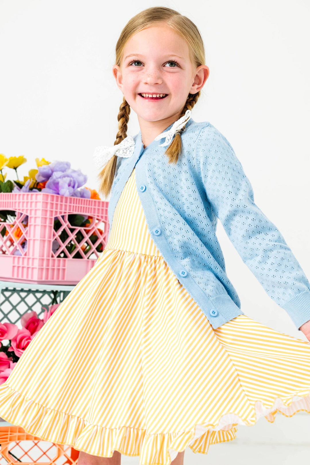 little girl twirling in front of flower stand in pastel yellow and white pinstripe twirl dress paired with spring blue pointelle knit cardigan