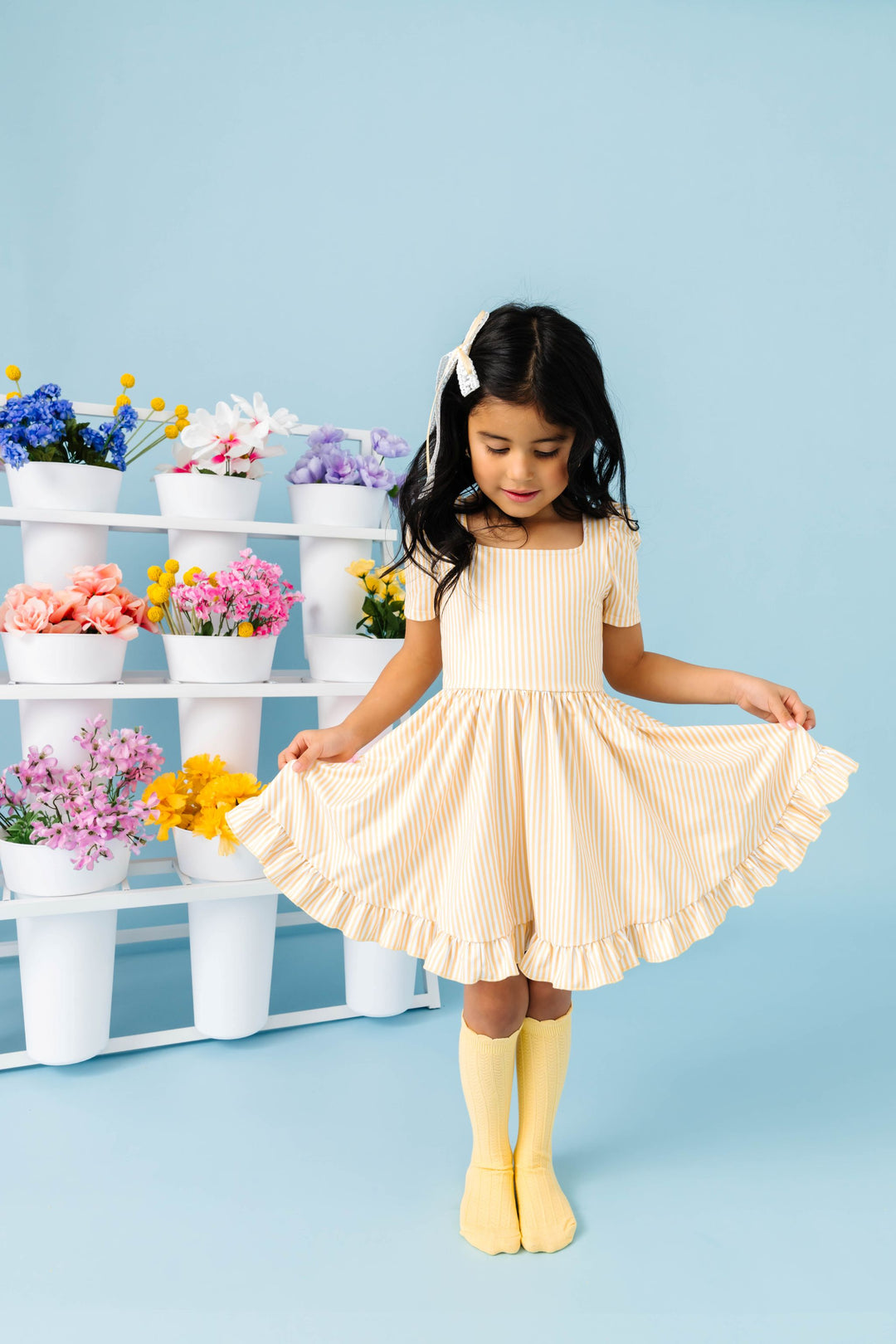 little girl in front of spring flower display wearing pastel yellow and white pinstripe twirl dress aired with yellow scalloped knee highs and matching lace ribbon bow