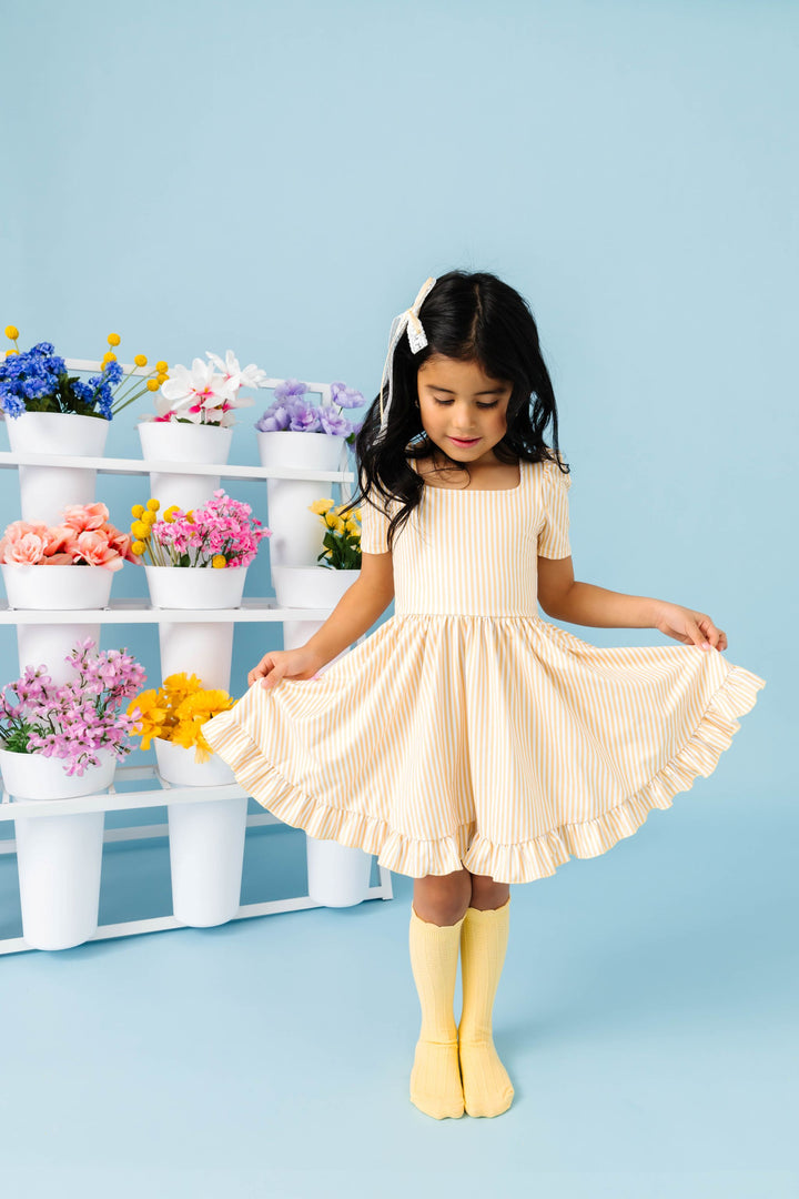 little girl in front of spring flower display wearing pastel yellow and white pinstripe twirl dress aired with yellow scalloped knee highs and matching lace ribbon bow