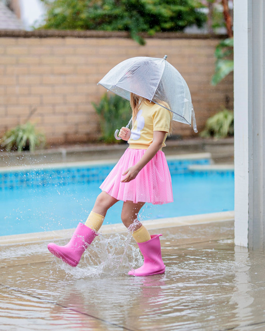 little girl splashing in rain puddle with umbrella wearing pastel yellow short sleeve sweater, pink tutu skort with white dots, yellow knee highs and pink rain boots