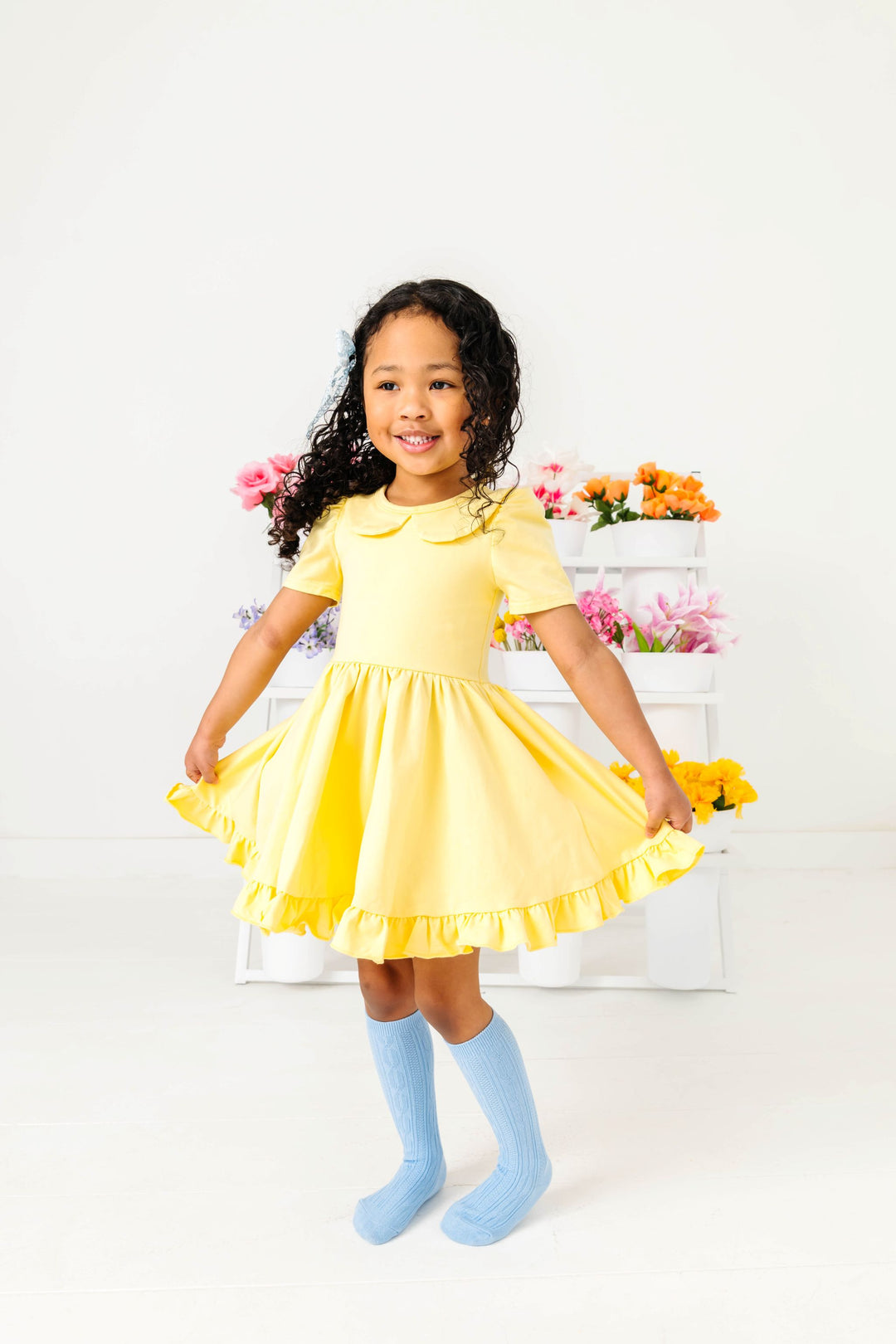 little girl twirling in front of flower display in pastel yellow cotton blend twirl dress with peter pan collar and light blue cable knit knee high socks