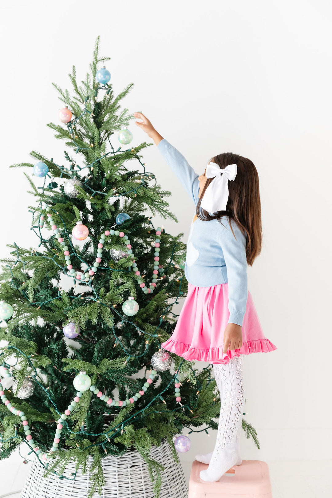 girl in front of tree wearing white satin hair bow, blue sweater, pink dress and white tights