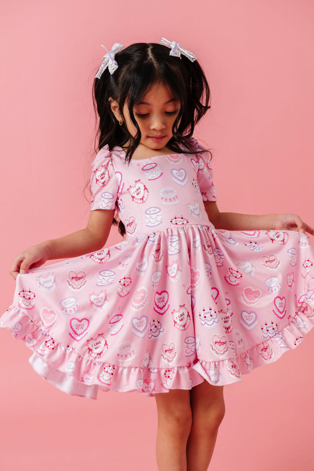 girl in light pink square neck twirl dress with sweet valentines cakes and words of affirmation paired with matching white lace pigtail bows with lavender ribbon
